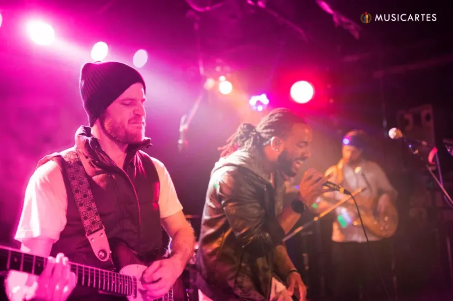 Banda tocando en vivo bajo luces rosadas: un guitarrista con gorro y un cantante.