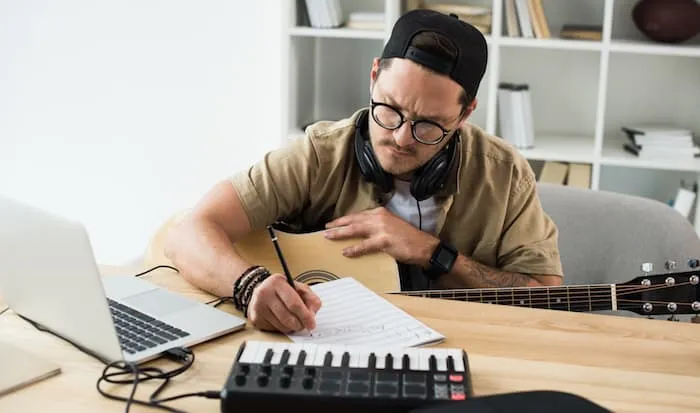 Músico con guitarra acústica escribiendo en una partitura frente a una computadora portátil y un teclado MIDI.