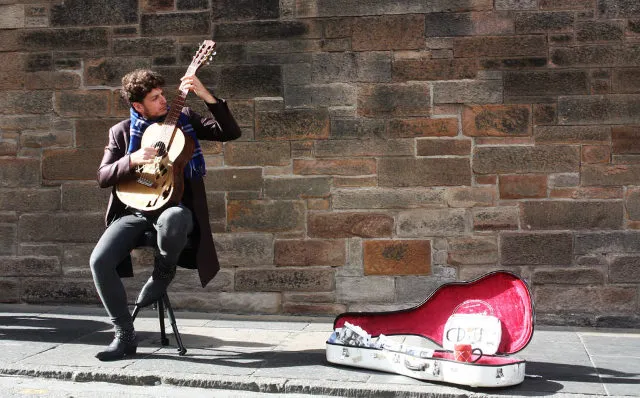 Músico callejero tocando guitarra junto a una pared de ladrillo, con el estuche abierto en la acera.